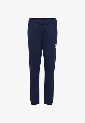 Joven de pie, con una camiseta gris de Hummel, pantalones azul marino, zapatillas blancas, con el pelo largo trenzado sobre un hombro, frente a un fondo liso.