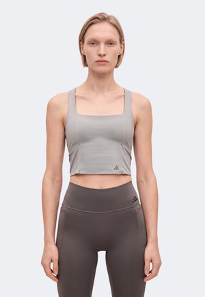 Une femme se tient face à l’objectif, vêtue d’un haut de sport court gris et d’un legging foncé, sur un fond clair uni.