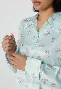 Himmelblaue Satinbluse mit pinkem Blumenmuster, ausgestattet mit einem spitzen Kragen, einer Knopfleiste und langen Ärmeln mit maßgeschneiderten Manschetten.