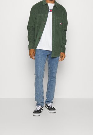 Mies, jolla on vihreä ribbineulospaita valkoisen graafisen t-paidan päällä, vaaleansiniset farkut ja mustavalkoiset tennarit, seisomassa yksinkertaisen taustan edessä.