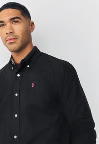 Črna bombažna srajca z gumbi, na prsih je rožnato izvezen logotip jastoga in na sprednji strani beli gumbi.