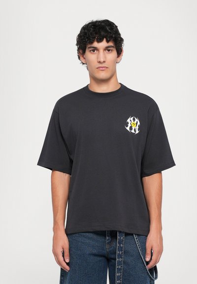 Camiseta negra de gran tamaño hecha de algodón, con un gráfico colorido en forma de llama en blanco y amarillo en el lado izquierdo del pecho. Mangas cortas.