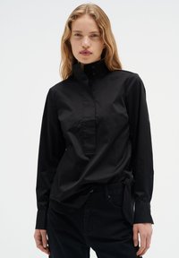 Schwarzes Button-Up-Hemd aus glattem Stoff, mit hohem Kragen und langen Ärmeln, leicht lockerer Passform und seitlichen Schlitzen.