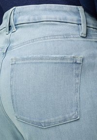 Κοντινό πλάνο στην πίσω τσέπη και θηλιά ζώνης από ανοιχτό μπλε τζιν σε άτομο που φοράει σκουρόχρωμο τοπ.