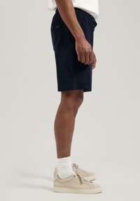 Marinen blå frottéshorts med elastisk midje, sidelommer og en avslappet passform, kombinert med beige sneakers og hvite sokker.