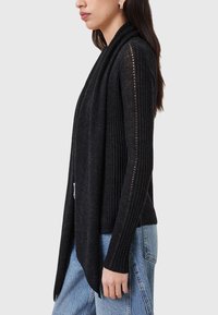 Black ribbed long-sleeve sweater with a draped front, open sides, and horizontal detailing. Paired with light blue denim jeans.
