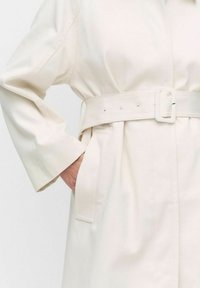 Beżowy płaszcz do kolan z paskiem w talii, długimi rękawami, kieszeniami bocznymi i gładką teksturą. Posiada kwadratową klamrę i minimalistyczny design.