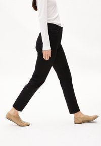 Jean noir coupe slim, design à cinq poches avec des détails de couture subtils, associé à des chaussures plates beiges. Texture de tissu lisse.