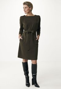 Olivgrön klänning med långa ärmar, knytning i midjan och sidofickor, tillsammans med eleganta svarta knähöga stövlar. Slät tygkvalitet, minimalistisk design.