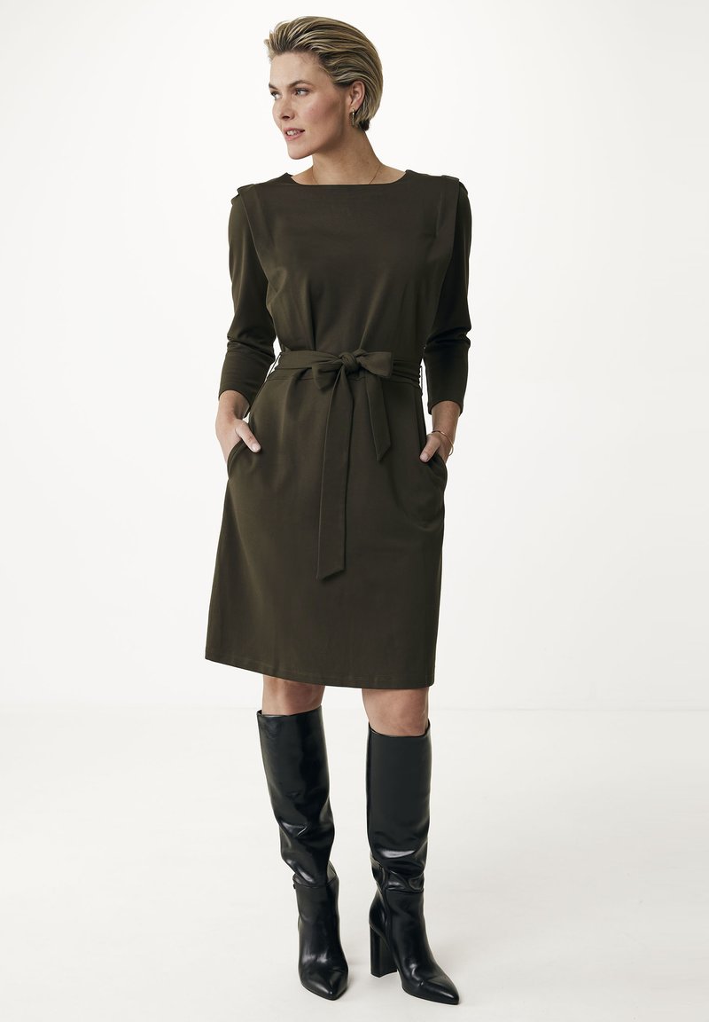 Oliwkowa sukienka z długimi rękawami, paskiem w talii i bocznymi kieszeniami, w połączeniu z eleganckimi czarnymi, sięgającymi kolan kozakami. Gładki materiał, minimalistyczny design.