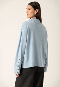 Light blue oversized sweater with a collar, featuring button accents along the sleeves, made from a soft fabric. Worn with dark pants.
