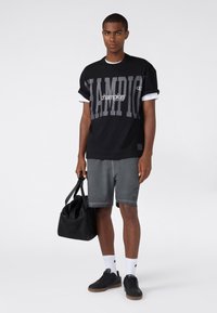 Giovane con una maglietta nera della Champion e pantaloni corti grigi, che tiene una borsa a tracolla nera, in piedi di fronte a uno sfondo chiaro e semplice.