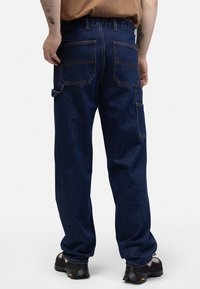 Pantalon en denim bleu foncé à coupe ample, avec des poches arrière doubles, une poche latérale pour outils et des détails de surpiqûres contrastées.