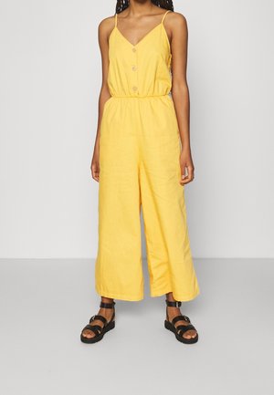 Kvinde iført en ærmeløs gul jumpsuit med knapper foran og elastisk talje, kombineret med sorte ankelstroppesandaler på en ensfarvet baggrund.