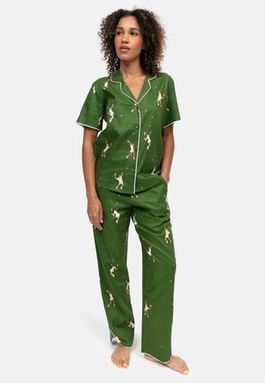 Femme portant un pyjama vert à manches courtes avec des illustrations de joueuses de tennis et une bordure blanche, debout pieds nus les mains dans les poches.