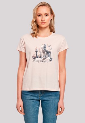 Light pink cotton t-shirt featuring a graphic print of a skeleton writing at a desk with candles and paper elements. Short sleeves, relaxed fit.