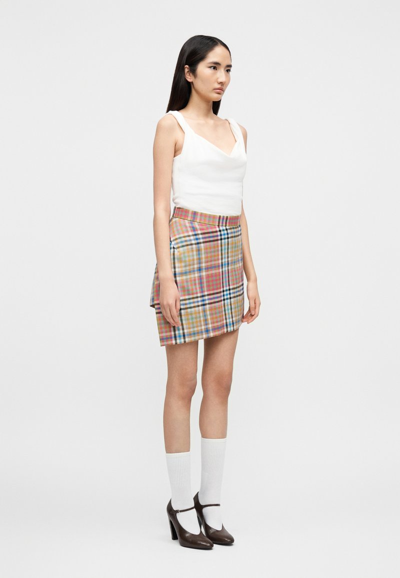 Frau trägt ein weißes, ärmelloses Oberteil, einen mehrfarbigen karierten Minirock, weiße Kniestrümpfe und braune Mary-Jane-Schuhe mit hohem Absatz.