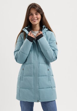 Eine lächelnde Frau trägt einen hellblauen, gepolsterten Wintermantel mit Kapuze und schwarzen Innenbündchen, hält den Kragen und steht vor einem weißen Hintergrund.