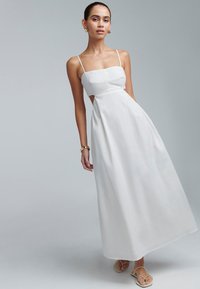 Vestido maxi blanco con tirantes finos, detalles cortados en la cintura y un dobladillo fluido. Tejido ligero con una textura suave.