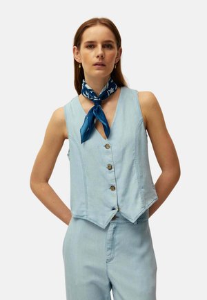 Mujer que lleva un chaleco sin mangas azul claro abotonado y pantalones a juego con un pañuelo azul con estampado alrededor del cuello, sobre un fondo liso.