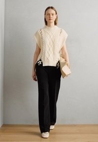 Kremfarget kabelstrikket oversized genser med sidesnører, kombinert med svarte bukser og en beige veske, satt mot en nøytral bakgrunn.