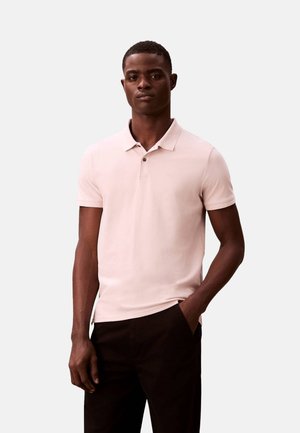 Joven con una camisa polo de manga corta color rosa claro y pantalones negros, de pie con una mano en el bolsillo contra un fondo liso.