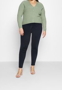 Cardigan côtelé vert clair avec col en V et boutons, associé à un jean skinny bleu foncé et des sandales à talons rose clair.