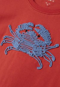 Camiseta de tela roja con un detallado diseño de cangrejo bordado en azul y ojos negros, que muestra textura y contraste.