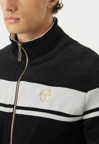 Svart glidelåsjakke med høy krage, med grå horisontale striper og en gulllogo. Laget av mykt materiale med en teksturert finish.