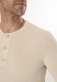 Beige gerippter Strickpullover mit Rundhalsausschnitt, Drei-Knopf-Leiste und strukturiertem Stoff. Sichtbare Nähte entlang der Raglanärmel.
