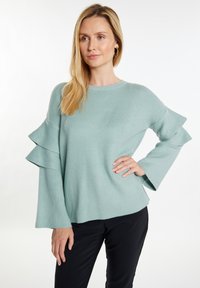 Šviesiai žalias megztinis su garbanotomis rankovių detalėmis. Turi apvalų iškirpimą ir laisvą siluetą, derinamas su juodomis kelnėmis. Lygus paviršius.