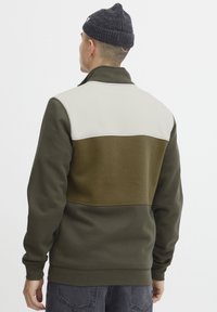 Giacca in pile con un design a blocchi di colore verde oliva e beige, colletto alto e chiusura con zip, realizzata in un tessuto morbido e strutturato.