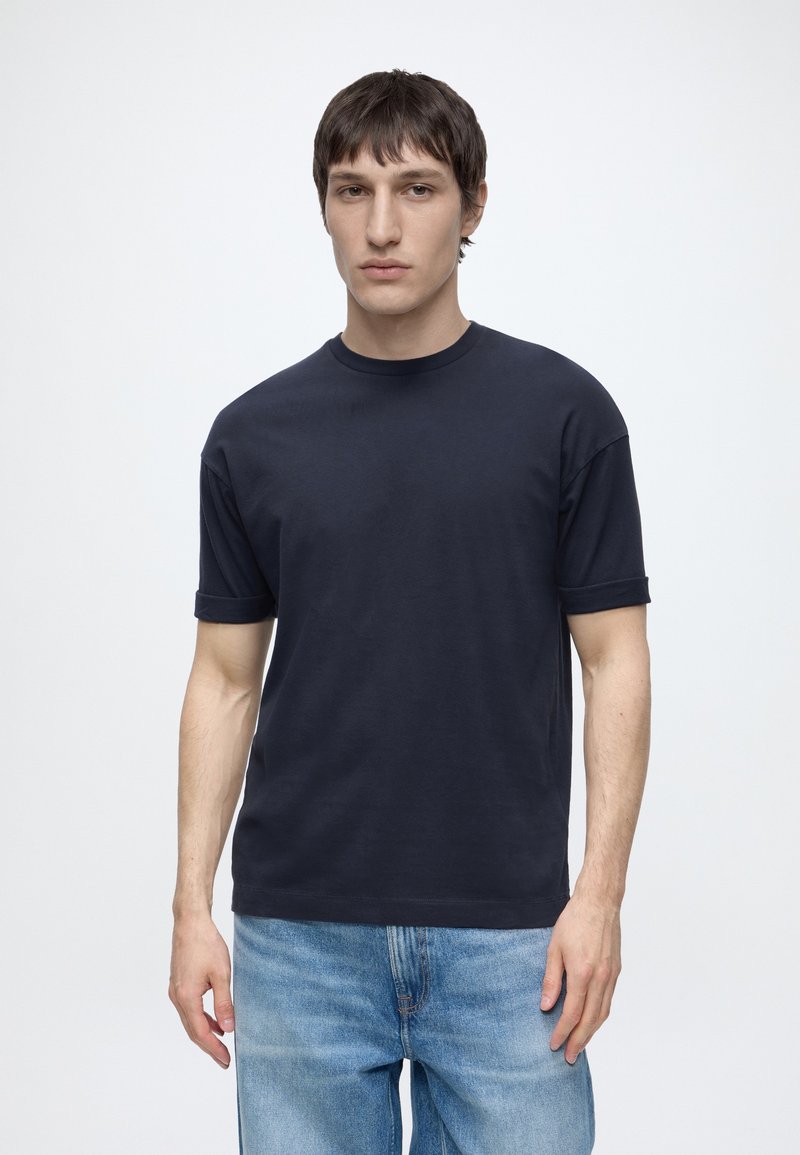 Jeune homme aux cheveux courts et foncés, portant un t-shirt noir uni et un jean bleu clair, debout sur un fond blanc.