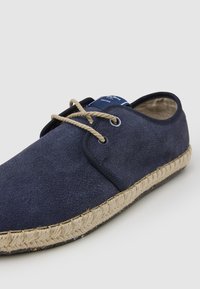 Zapatos de ante azul marino tipo slip-on con suela de cuerda beige, que cuentan con dos ojales y cordones de color marrón claro, ofreciendo un diseño texturizado y casual.