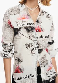 Witte button-up shirt met een bloem- en vlinderprint, gemengd met tekst- en krantenmotieven. Lange mouwen met opgerolde manchetten.