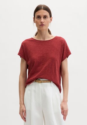 Femme aux cheveux foncés attachés en queue de cheval basse, portant un haut rouge à manches courtes et ample, rentré dans un pantalon blanc taille haute avec une ceinture beige.