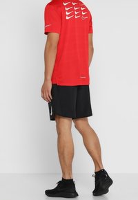 Camiseta atlética roja de Nike con acentos de logo blancos, combinada con pantalones cortos negros y calzado deportivo negro. Vista desde atrás.