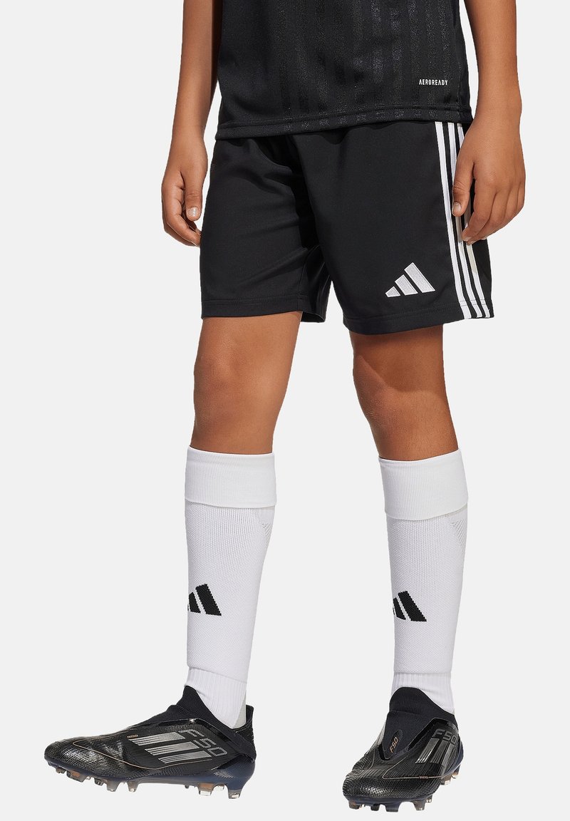 Pantalones cortos deportivos negros con acentos de tres franjas blancas, combinados con calcetas blancas hasta la rodilla. Botas de fútbol negras con diseño texturizado y tachuelas.