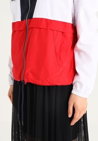 Czerwono-biała kurtka przeciwwiatrowa z czarnym paskiem, z zapięciem na zamek, elastycznymi mankietami i przednią kieszenią. Nosi się ją na czarną spódnicę plisowaną.