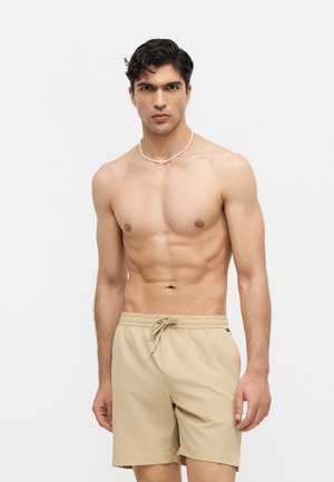 Μυώδης άνδρας με σκούρα μαλλιά, ντυμένος με μπεζ μαγιό βερμούδα και λευκό κολιέ, στέκεται μπροστά σε απλό ανοιχτό φόντο.