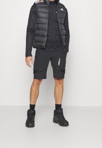 Muž v černé péřové vestě Adidas, tričku s dlouhým rukávem, kraťasech Terrex, černých ponožkách a černých turistických botách stojí na jednobarevném pozadí.