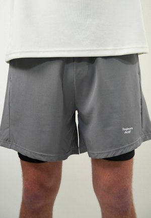 Homme portant un short de sport gris par-dessus un short de compression noir et un t-shirt blanc, montrant le logo "Toujours Actif" sur le short gris.