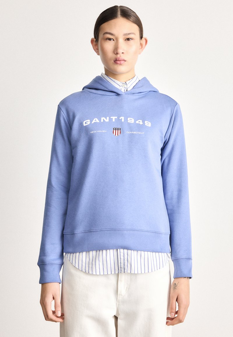 Sudadera azul claro con capucha que presenta el texto "GANT1949" y un logo. El diseño incluye un dobladillo y puños acanalados, superpuestos sobre una camisa a rayas.