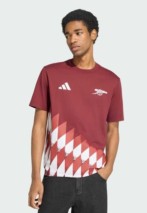 Jeune homme portant un maillot de football Arsenal bordeaux avec des logos blancs adidas et canon, ainsi qu’un motif en losange blanc et rouge sur la moitié inférieure.