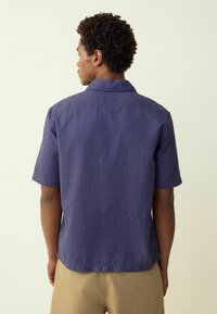 Short-sleeve shirt in textured dark blue fabric, with a collar and open back design, featuring small, raised patterns throughout.