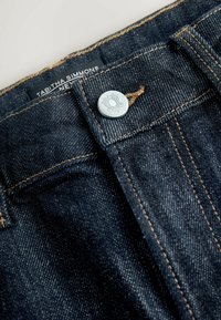 Jeans de denim oscuro con cierre de botón plateado, costuras en contraste en color naranja y una etiqueta tejida que dice "Tabitha Simmons" en la cintura.