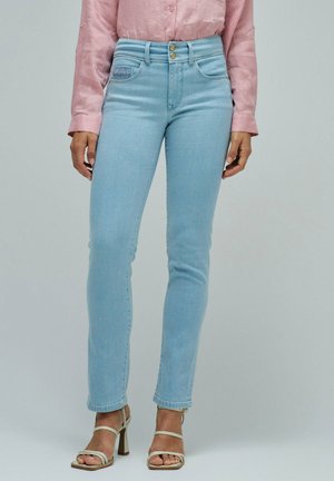 Mujer que lleva vaqueros pitillo de tiro alto azul claro, blusa rosa de manga larga y sandalias de tacón beige, de pie frente a un fondo liso.