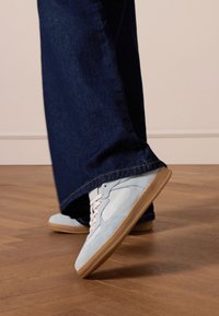 Lichtblauwe sneakers van suede en leer met witte veters, gecombineerd met donkerblauwe, wijde denim jeans. Achtergrond van houten vloer.