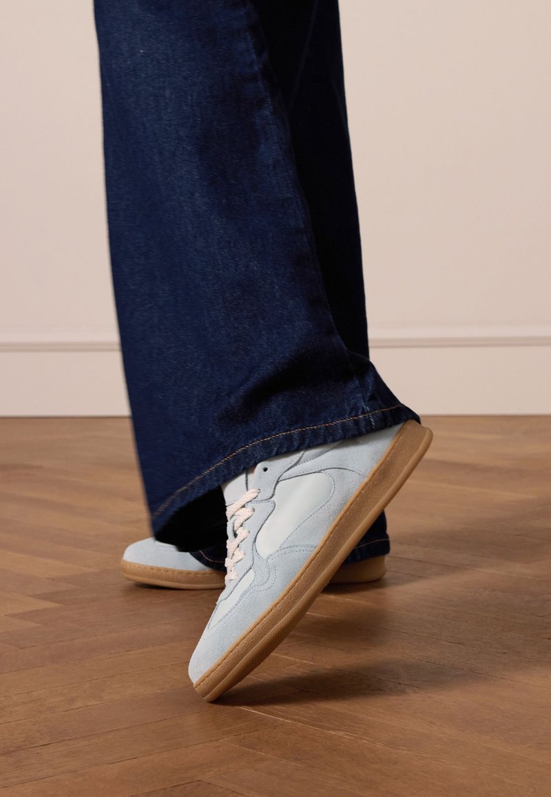 Light blue sneakers made of suede and leather with white laces, paired with dark blue, wide-leg denim jeans. Wood floor background.