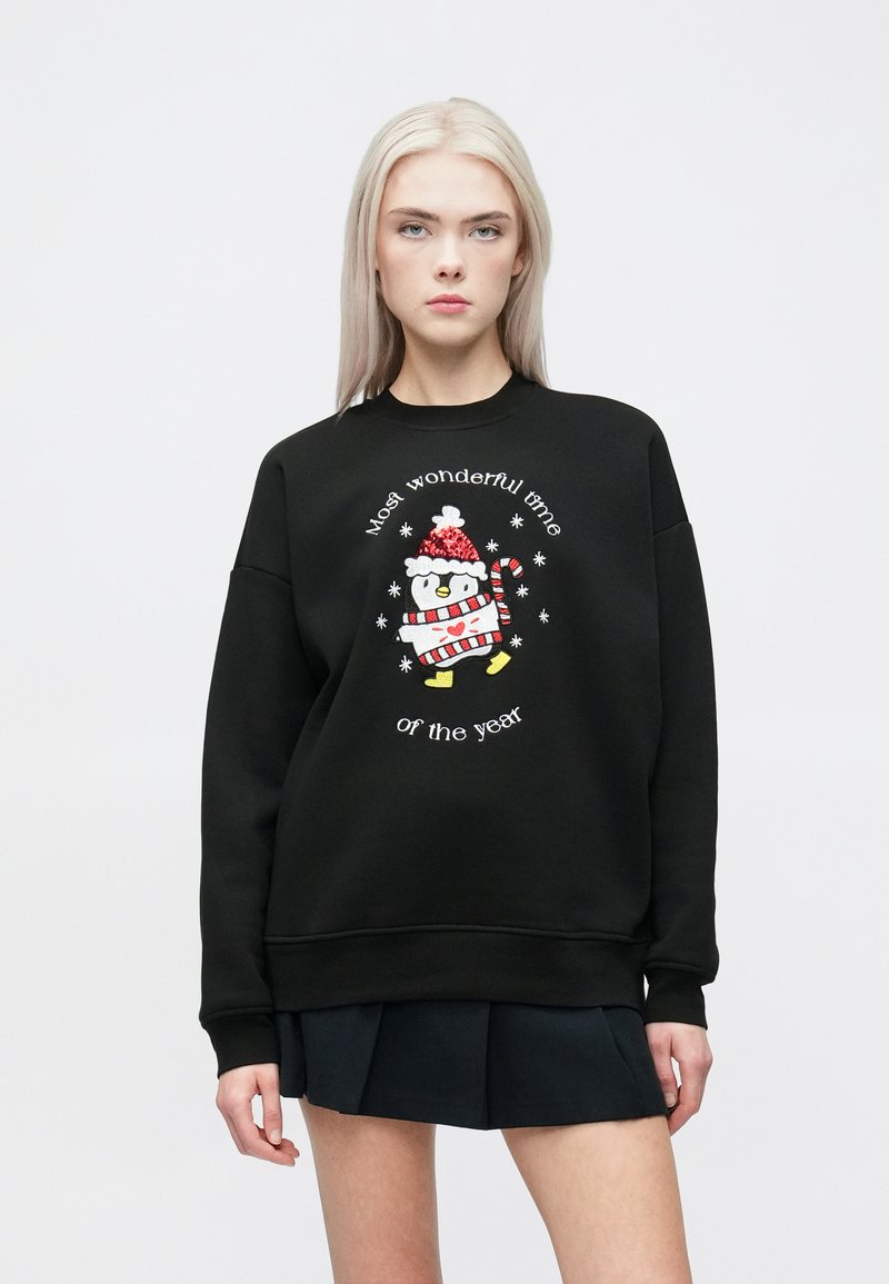 Melna sudraba jaka ar izšūtu pingvīnu, kuram virsū ir Ziemassvētku cepure un svītrainā krekls, ap to ir teksts "Brīnišķīgākais gada laiks".